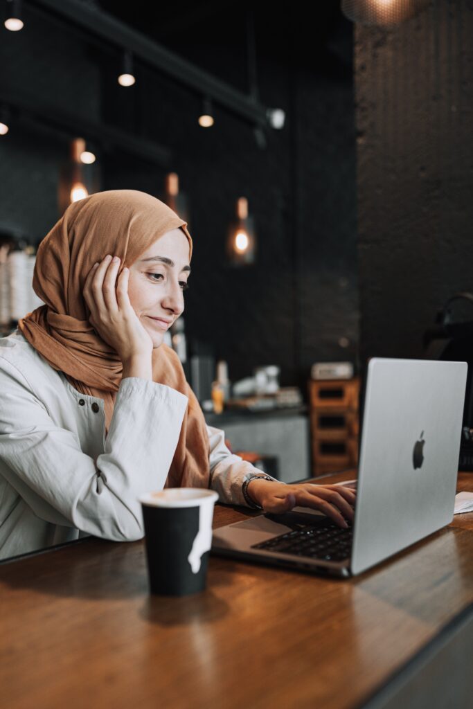 pexels-photo-17564434-17564434 A woman in a headscarf works on her laptop in a warm café setting, enjoying a coffee.